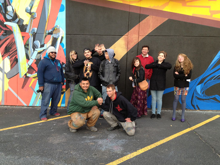 A group of ten young artists posing in front of the mural they have just finished. Two of them are kneeling and shaking hands.