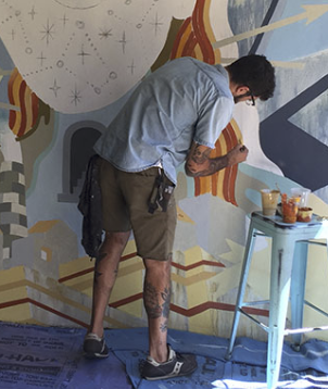A photo of the young tattooed artist in shorts with his back turned painting the mural in progress.