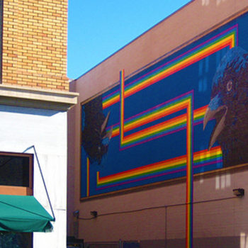 Photo in a small alley of a large mural with a stylized rainbow and two crow heads. One is upside-down.