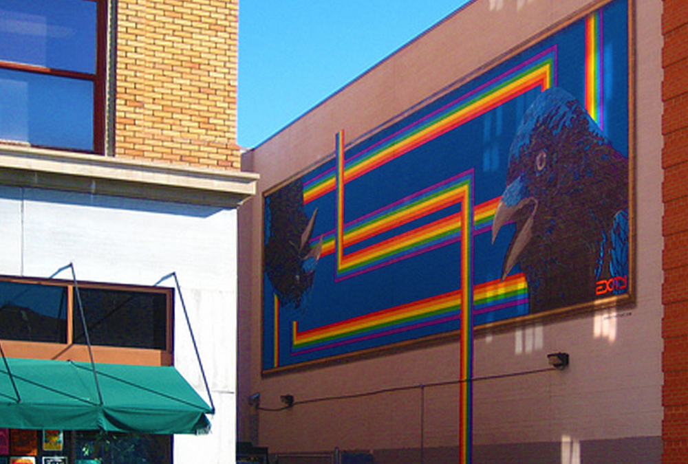 Photo in a small alley of a large mural with a stylized rainbow and two crow heads. One is upside-down.