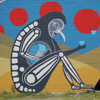 Photo of a mural of a strnge creature sitting down that is kind of a cross between a human sketeton body and a bird head with circles surrounding it.