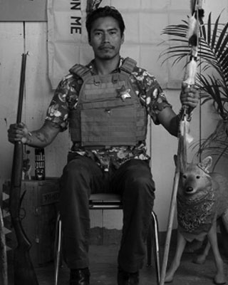man seated facing camera holding gun and stick